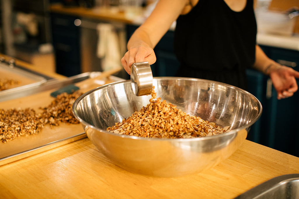 Mixing up a batch of gluten free oat granola in a large stainless steel bowl in a kitchen. Adding in pecan pieces.